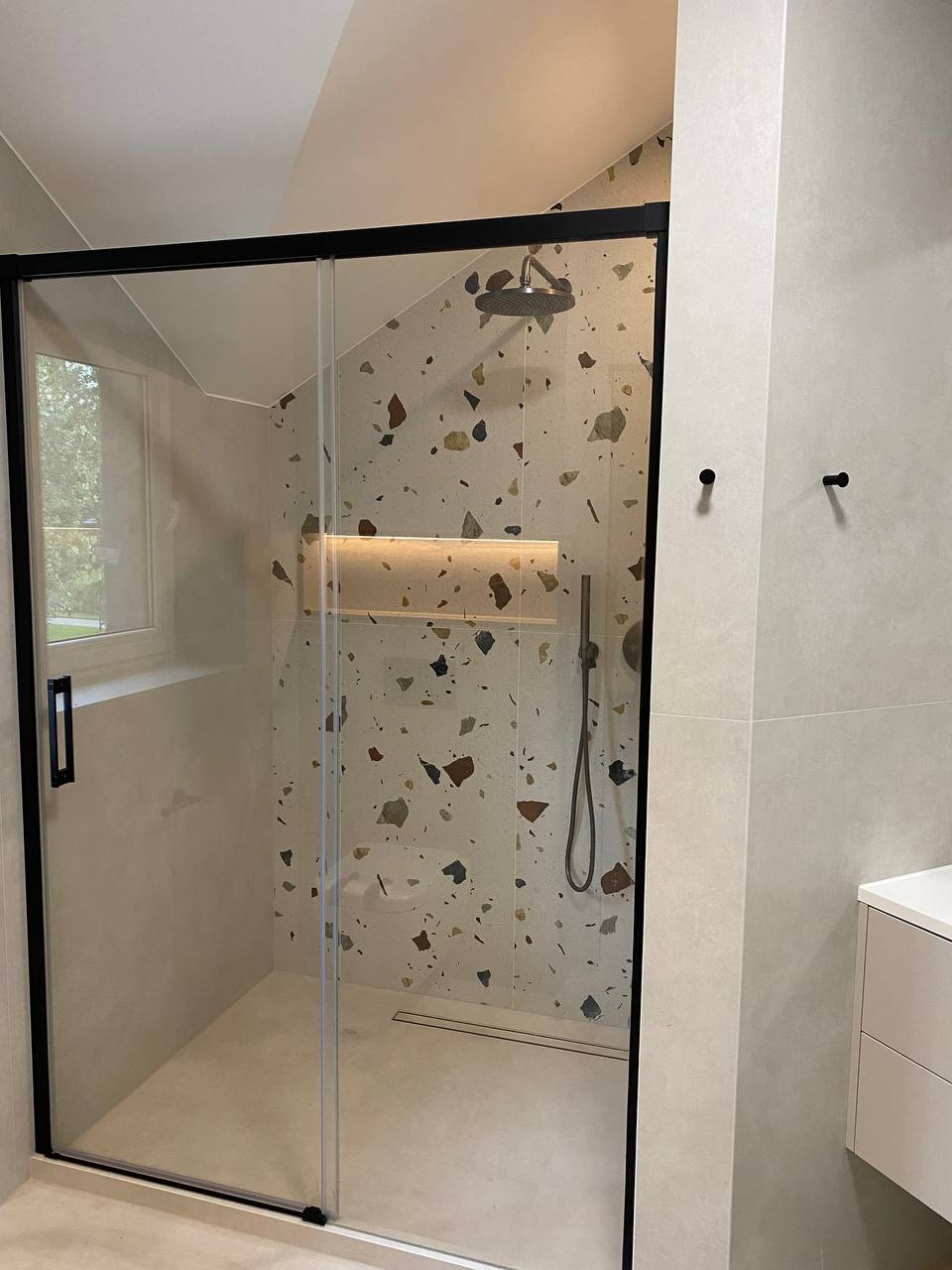 Nowoczesna kabina prysznicowa z czarnymi ramami i ścianą wyłożoną płytkami w stylu lastryko, widoczne czarne wieszaki na ręczniki i oświetlona wnęka prysznicowa.