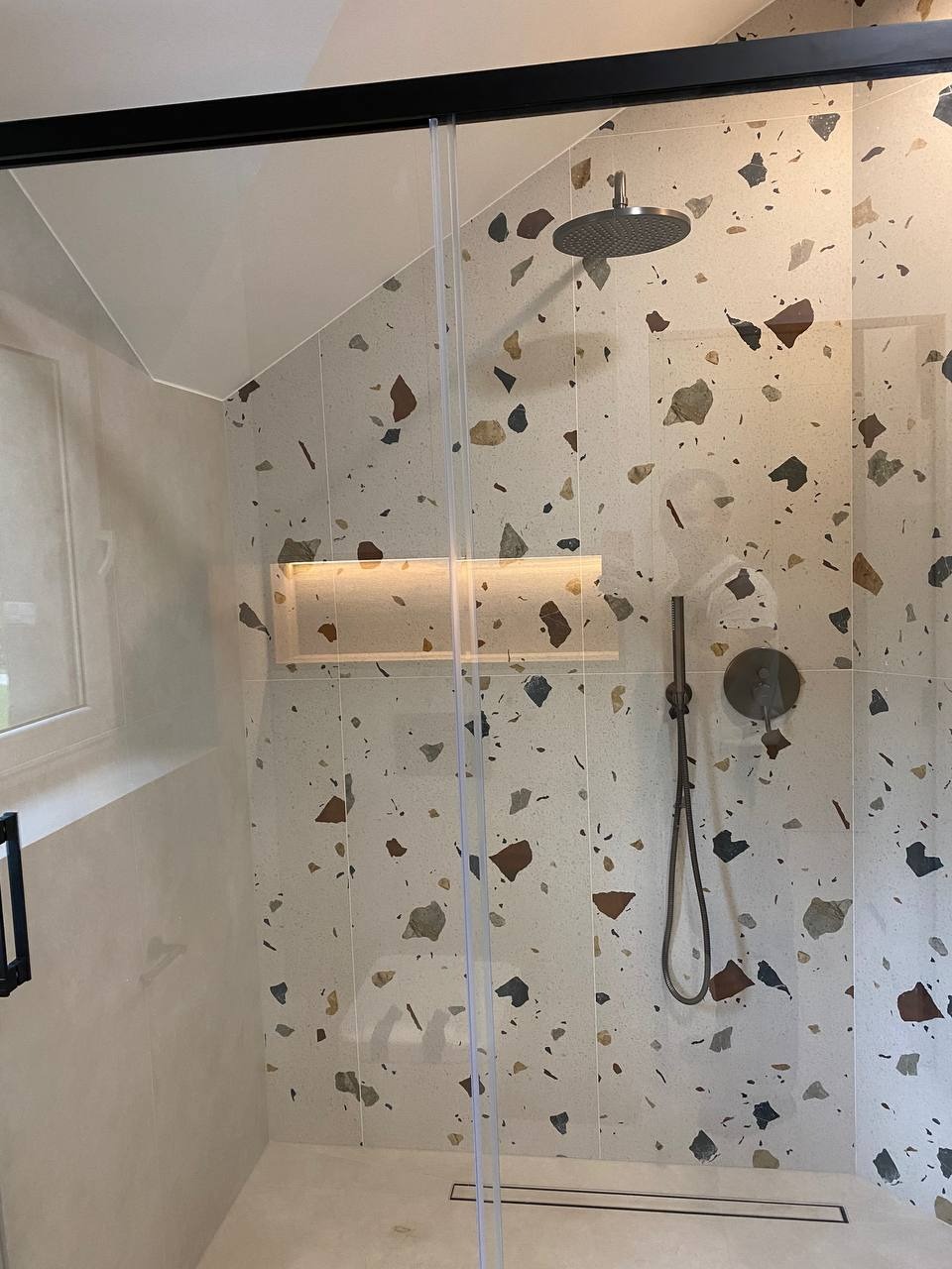 Nowoczesna kabina prysznicowa z deszczownicą i wnęką oświetleniową, wykończona płytkami w stylu lastryko z elementami brązowymi, szarymi i beżowymi.