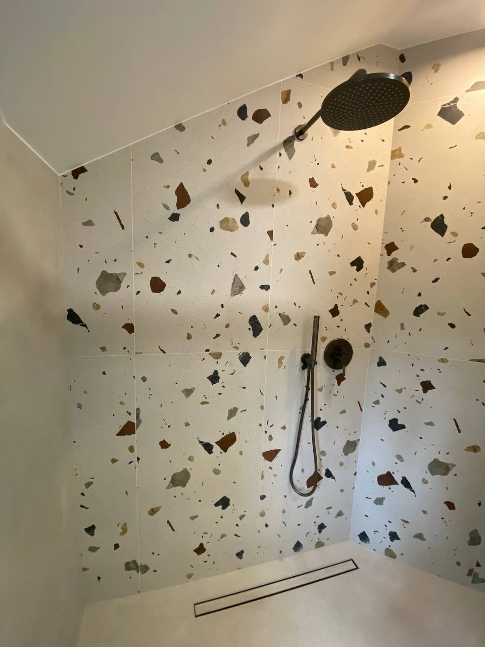 Kabina prysznicowa z wykończeniem ścian płytkami imitującymi lastryko w odcieniach brązu, szarości i czerni, z czarną deszczownicą i odpływem liniowym.