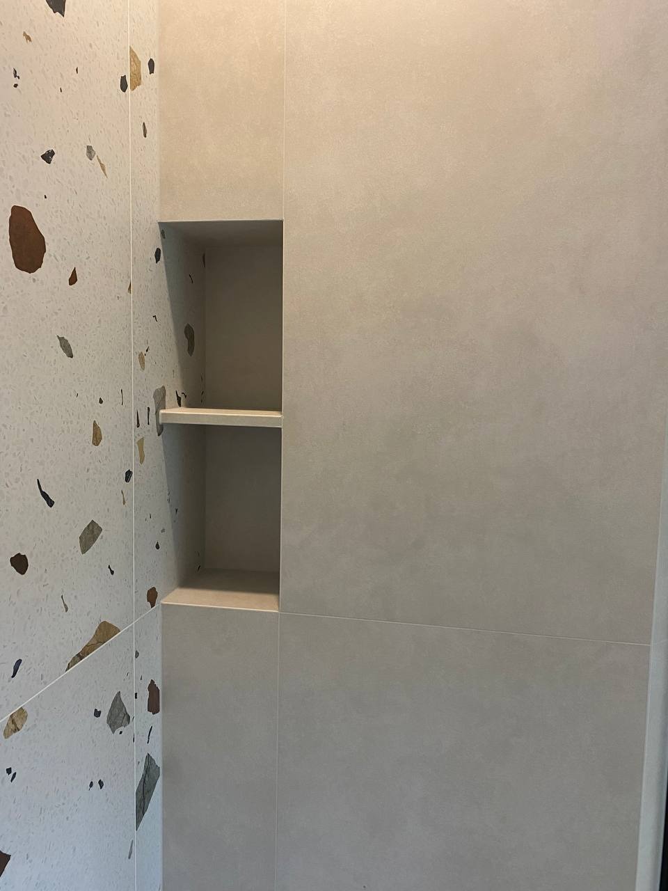 Wnęka z półkami w ścianie wyłożonej szarymi płytkami, obok ściana z białymi płytkami imitującymi lastryko.