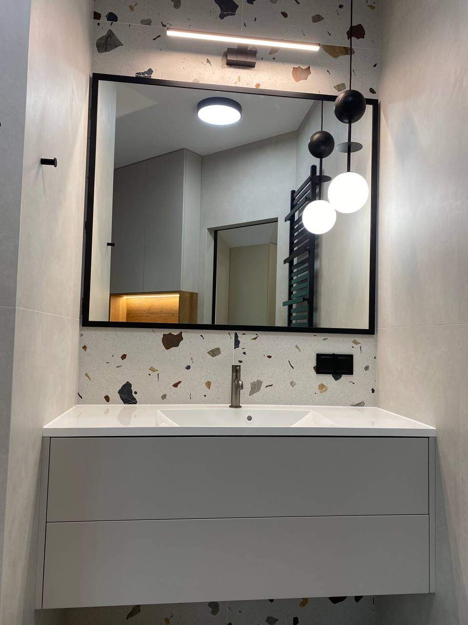 Nowoczesna łazienka z umywalką nablatową, szafką pod umywalką w odcieniach szarości, lustrem w czarnej ramie i mozaikową ścianą w tle.