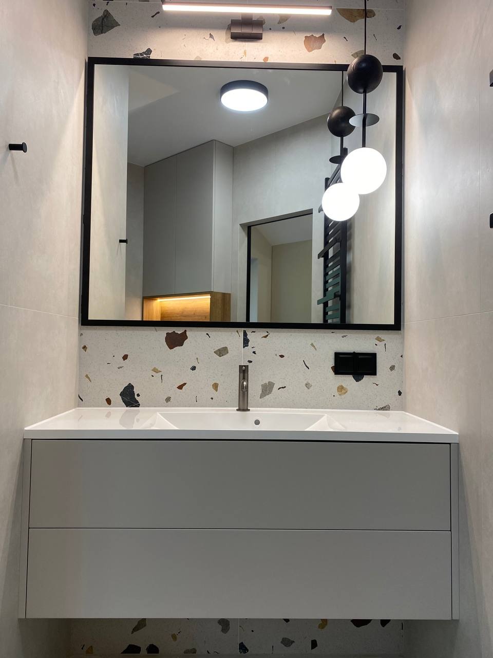 Nowoczesna łazienka z umywalką w szafce, lustrem w czarnej ramie i oświetleniem kulistym. Uwagę zwraca ściana i podłoga wykończona płytkami imitującymi lastryko.