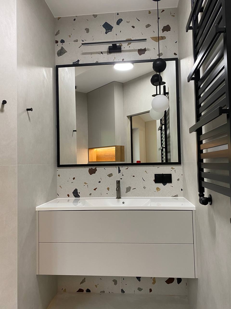 Nowoczesna łazienka z umywalką w szafce, lustrem w czarnej ramie, grzejnikiem drabinkowym i lampą wiszącą z kulami, płytki imitujące lastryko na ścianach i podłodze.