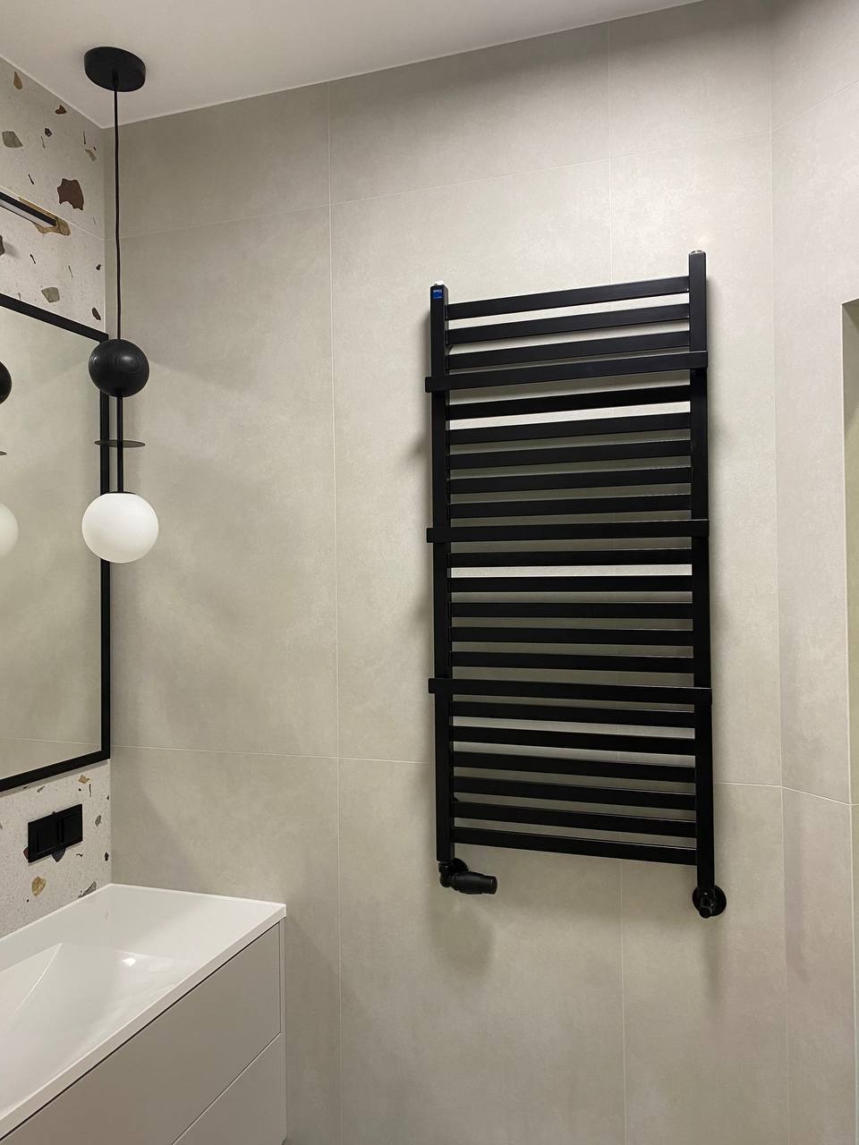 Nowoczesna łazienka z czarnym grzejnikiem drabinkowym na kremowych płytkach, minimalistyczna umywalka z szafką i designerska lampa wisząca z kulami.