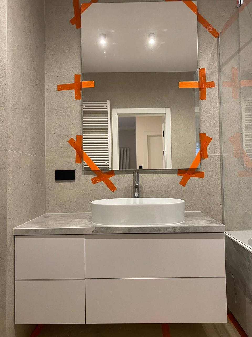 Nowoczesna łazienka w trakcie wykańczania: biała umywalka nablatowa na szarej szafce, lustro zabezpieczone pomarańczową taśmą, szare płytki na ścianach, widoczny grzejnik drabinkowy i fragment...