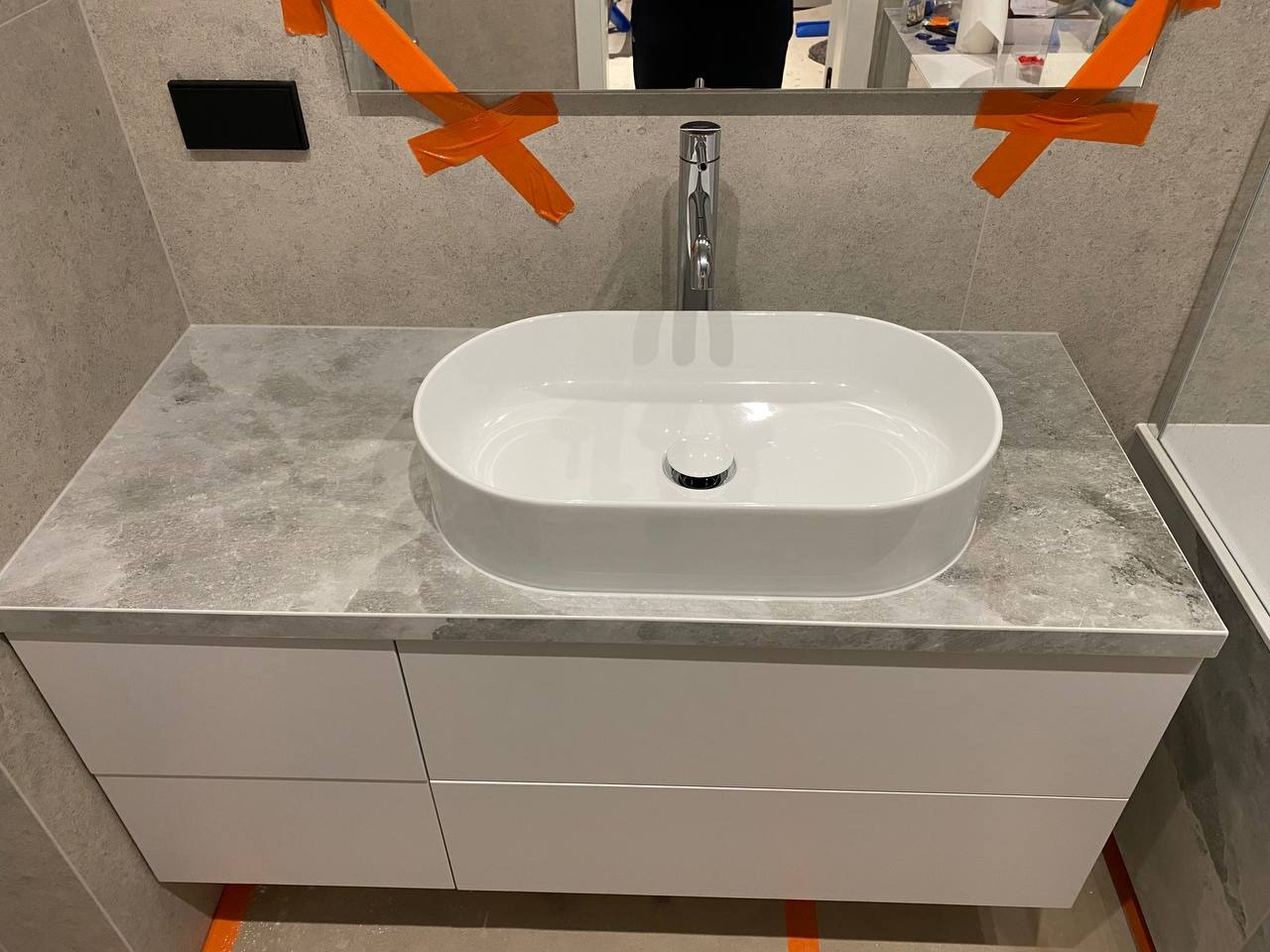 Owalna, biała umywalka nablatowa na szarym blacie z imitacją kamienia w łazience w Warszawie, widoczne taśmy zabezpieczające lustro.