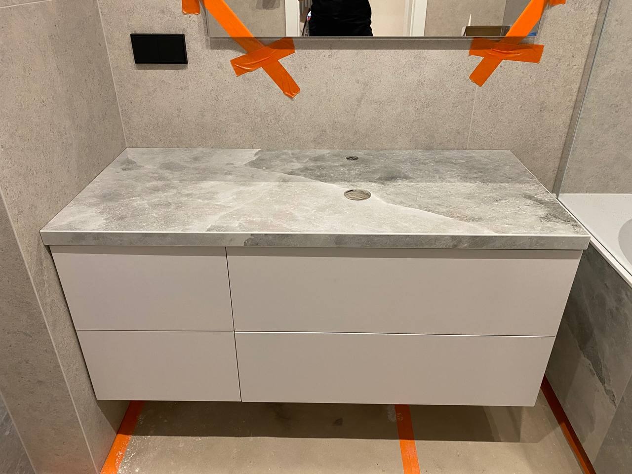 Montaż szafki pod umywalkę z szarym blatem imitującym marmur, widoczne taśmy zabezpieczające lustro i otwór na baterię, szare płytki na ścianach.
