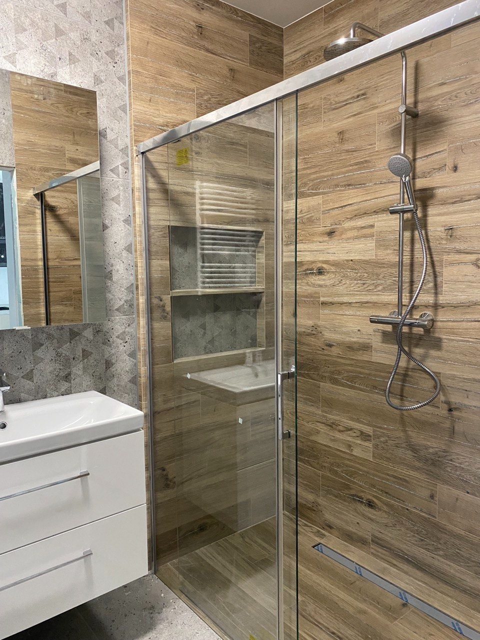 Nowoczesna łazienka z kabiną prysznicową typu walk-in, wykończona płytkami imitującymi drewno oraz szarą mozaiką, z białą szafką pod umywalką i chromowaną armaturą.