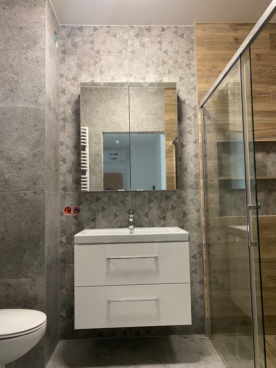 Nowoczesna łazienka z szaro-białymi płytkami w geometryczne wzory, białą szafką pod umywalką z chromowanymi uchwytami, lustrem z szafką i kabiną prysznicową z drewnopodobnymi panelami.