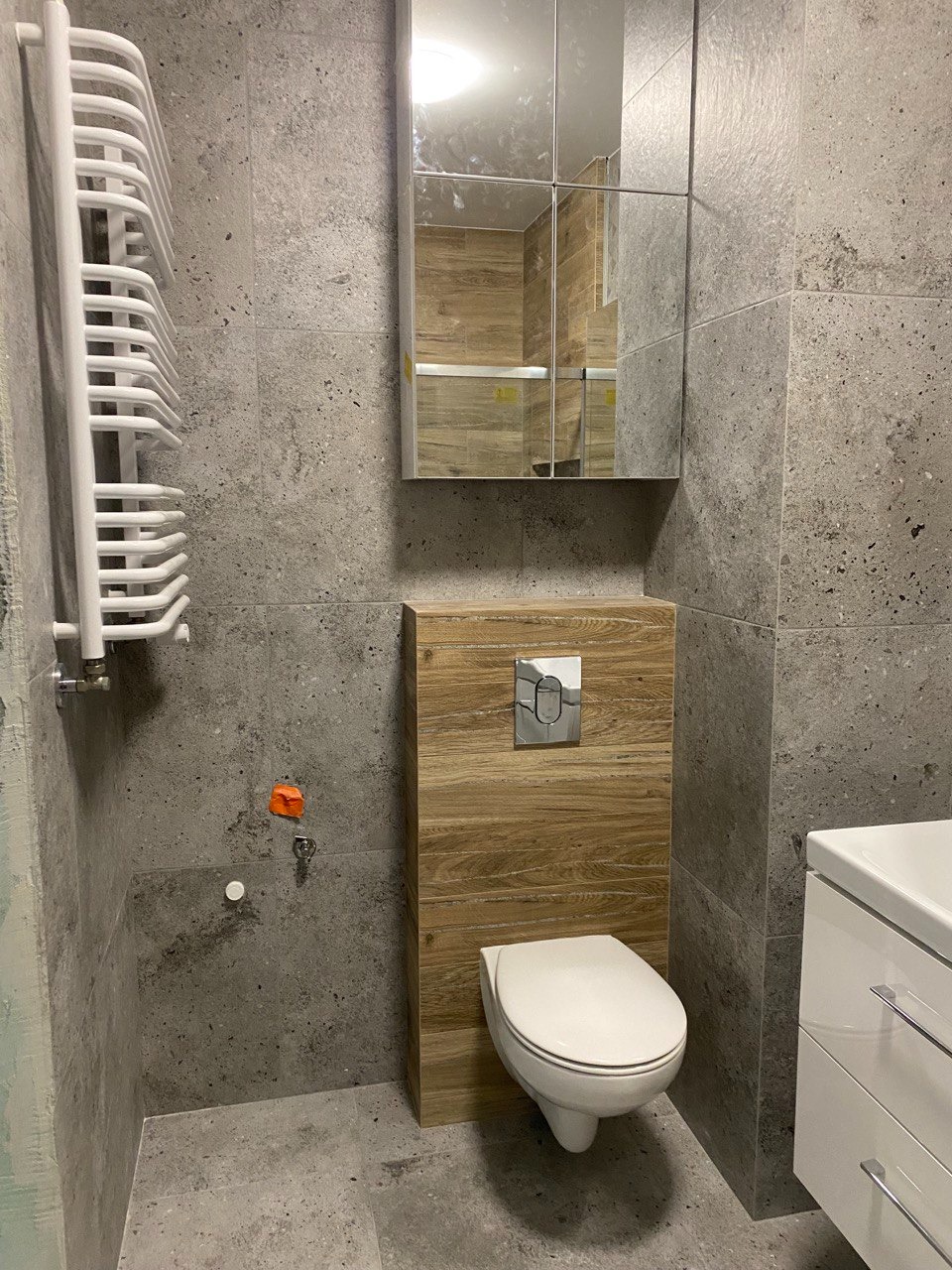 Nowoczesna łazienka z szarymi płytkami imitującymi kamień, wiszącą toaletą w zabudowie z drewnopodobnych paneli, lustrem szafkowym i białym grzejnikiem drabinkowym.