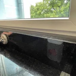 Montaż czarnego parapetu z połyskliwego kamienia z użyciem przyssawki, widok na okno i zieleń na zewnątrz, detale wykończenia.