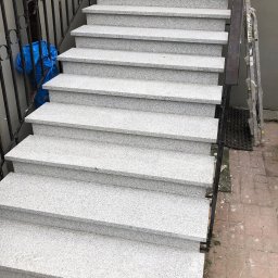 Kamień w Domu - Zewnętrzne schody z jasnego, nakrapianego kamienia z czarną metalową balustradą i drabiną opartą o ścianę w tle. Widok z góry na stopnie.