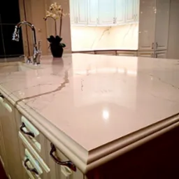 Błyszczący, kremowy blat z subtelnym marmurkowym wzorem na wyspie kuchennej z chromowaną armaturą i białymi szafkami w tle, oświetlony ciepłym światłem.