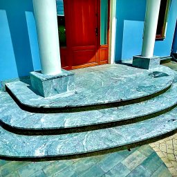 Kamień w Domu - Wejście do domu z trzema marmurowymi stopniami i kolumnami. Niebieska ściana, drewniane drzwi. Widok na schody i podjazd z kostki brukowej.