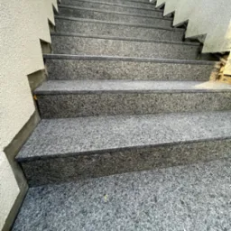 Zewnętrzne schody z szarego kamienia prowadzą w górę, widok z dołu. Ściana z szorstkim tynkiem po lewej stronie. Górne stopnie oświetlone słońcem.