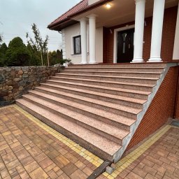 Kamień w Domu - Zewnętrzne schody z różowego kamienia prowadzące do domu z kolumnami, otoczone brukowaną ścieżką i kamiennym murem. Pochmurne niebo w tle.