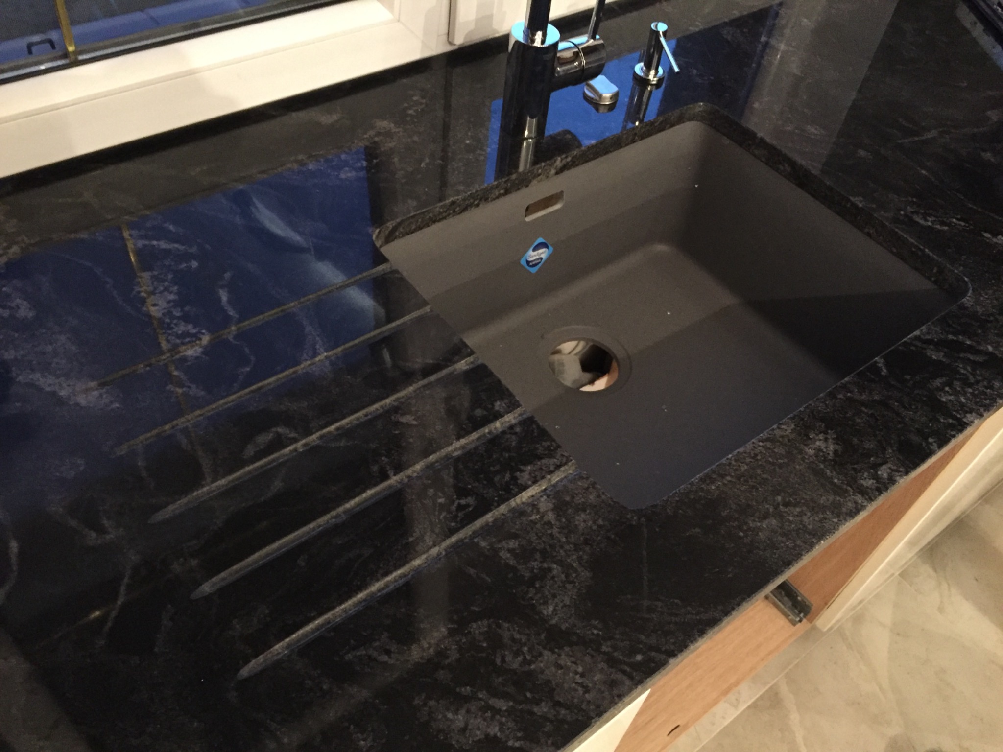 Czarny, polerowany blat z ciemnego kamienia z ociekaczem i wpuszczonym, kwadratowym, grafitowym zlewem oraz chromowaną baterią.