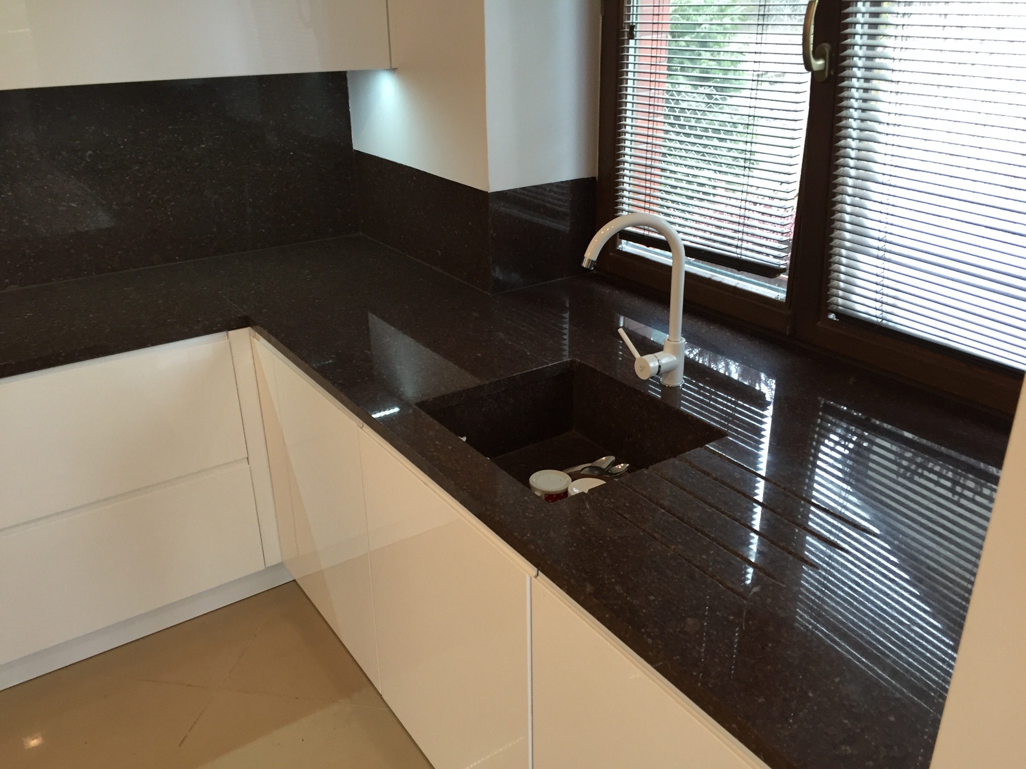 Ciemny, błyszczący blat kuchenny z wbudowanym zlewem i białą armaturą pod oknem z żaluzjami, uzupełniony białymi szafkami.