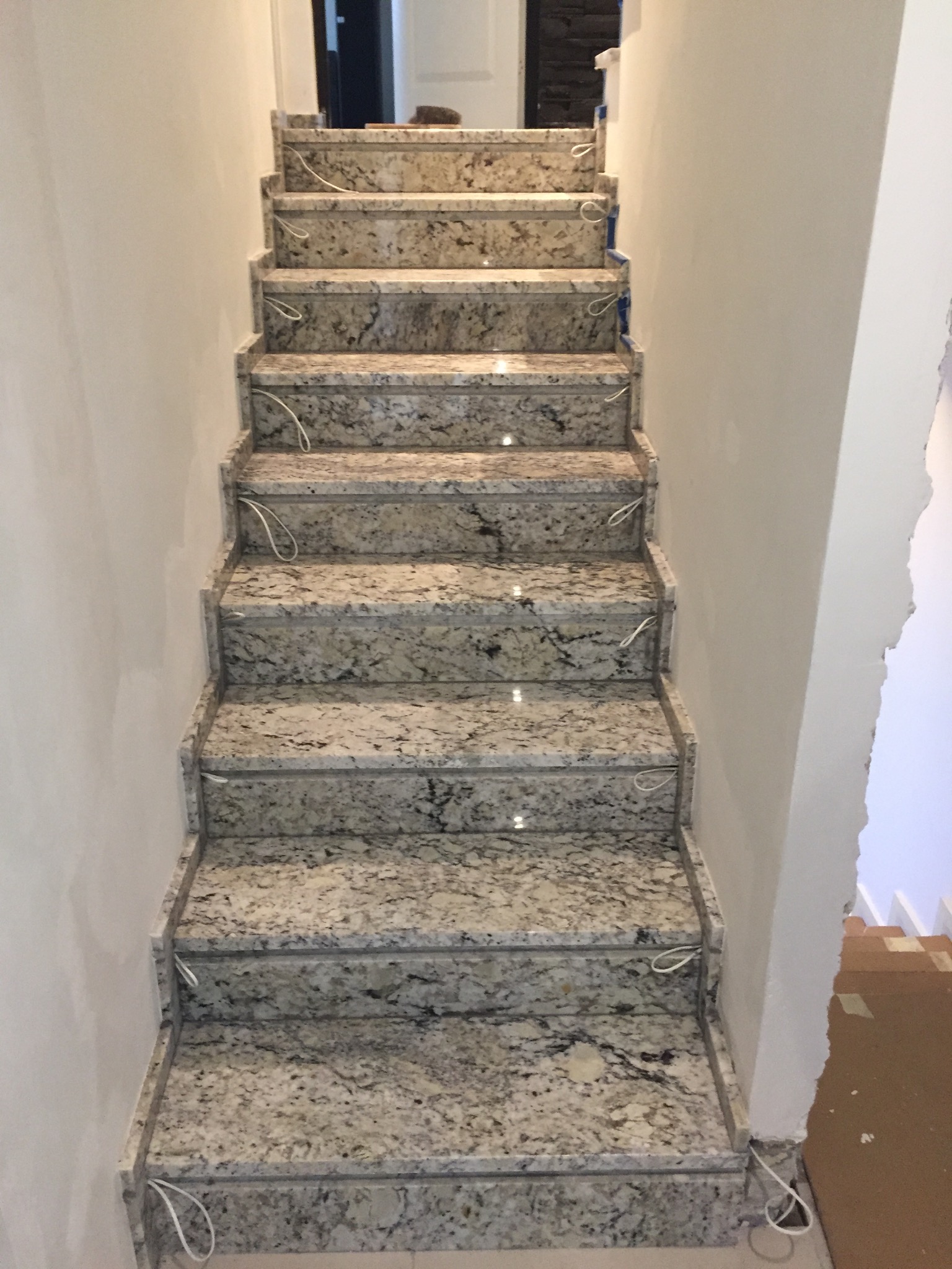 Schody wewnętrzne wyłożone jasnym kamieniem z ciemnymi plamkami, widoczne przewody elektryczne do oświetlenia ledowego na każdym stopniu, ściany w trakcie wykończenia.