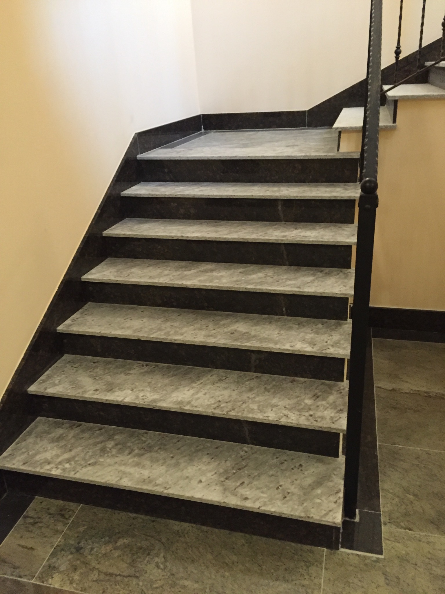 Wewnętrzne schody z szarymi stopniami i ciemną obudową, z czarną metalową balustradą, widoczne z dołu.