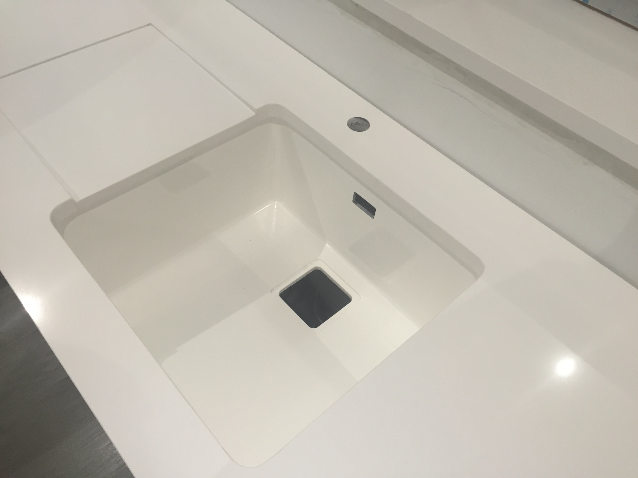 Biały, minimalistyczny blat kamienny z wbudowanym, kwadratowym zlewem i małym odpływem, sfotografowany z góry w jasnym oświetleniu.