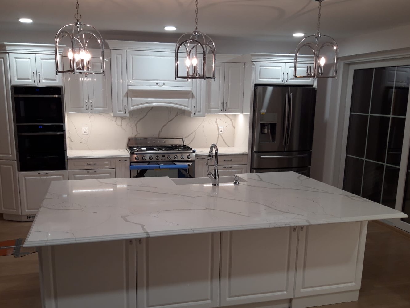 Biała kuchnia z wyspą i blatem z jasnego kamienia z szarymi żyłami, oświetlona trzema wiszącymi lampami w metalowych kloszach w kształcie klatek.