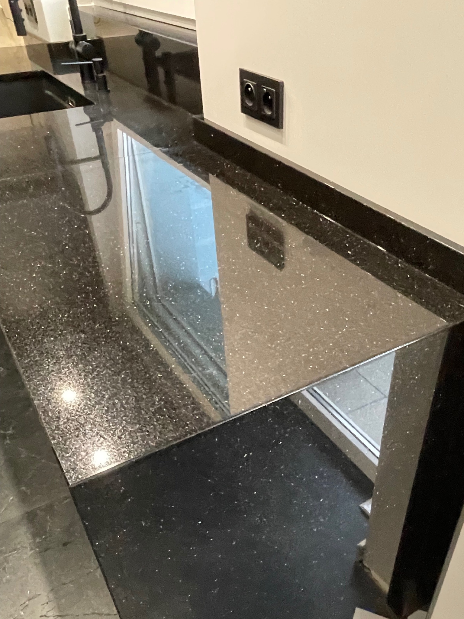 Błyszczący, czarny blat kamienny z brokatowym efektem, zamontowany przy oknie, z widocznym fragmentem zlewu i gniazdkiem elektrycznym na ścianie.