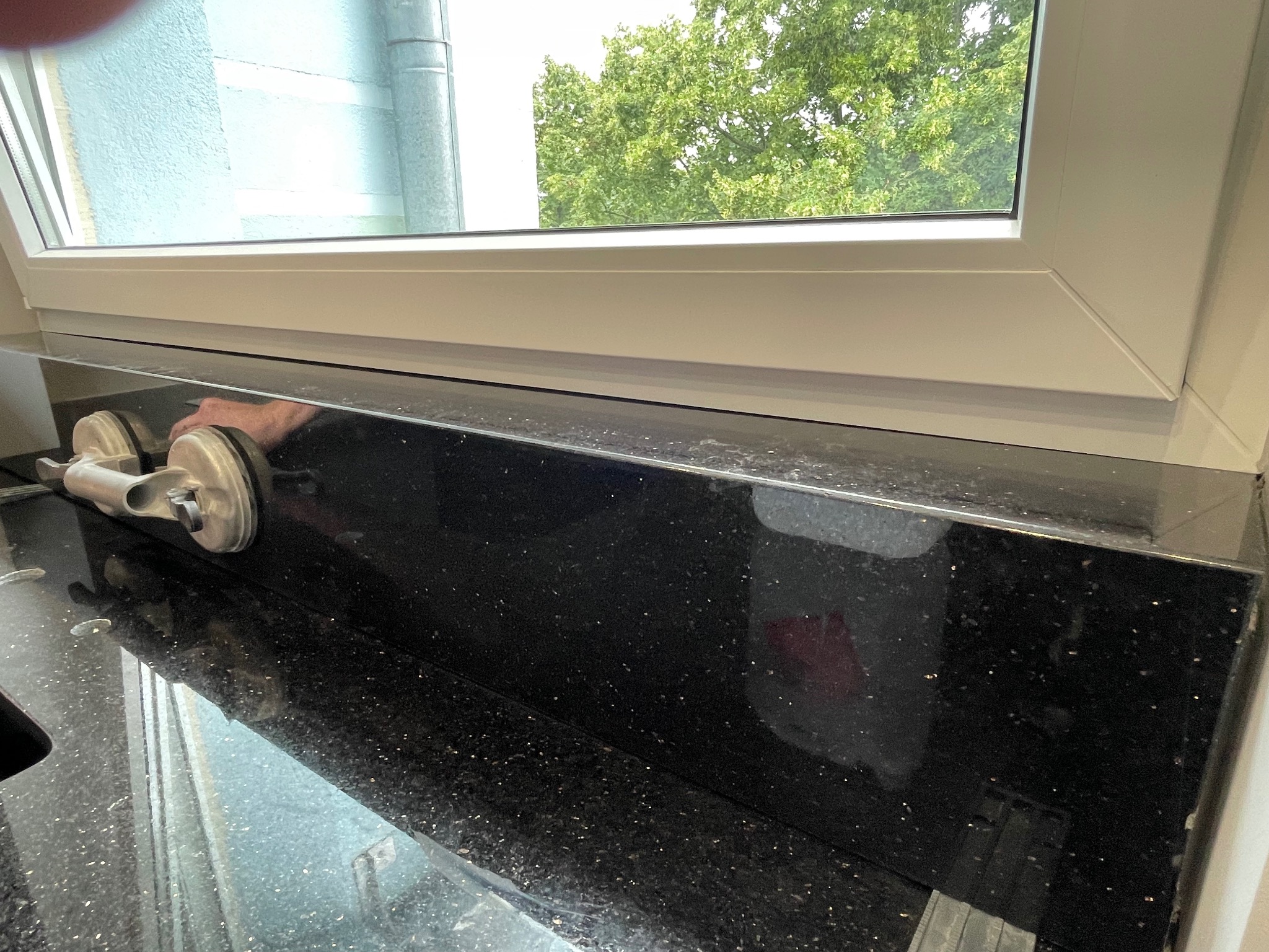 Montaż czarnego parapetu z połyskliwego kamienia z użyciem przyssawki, widok na okno i zieleń na zewnątrz, detale wykończenia.