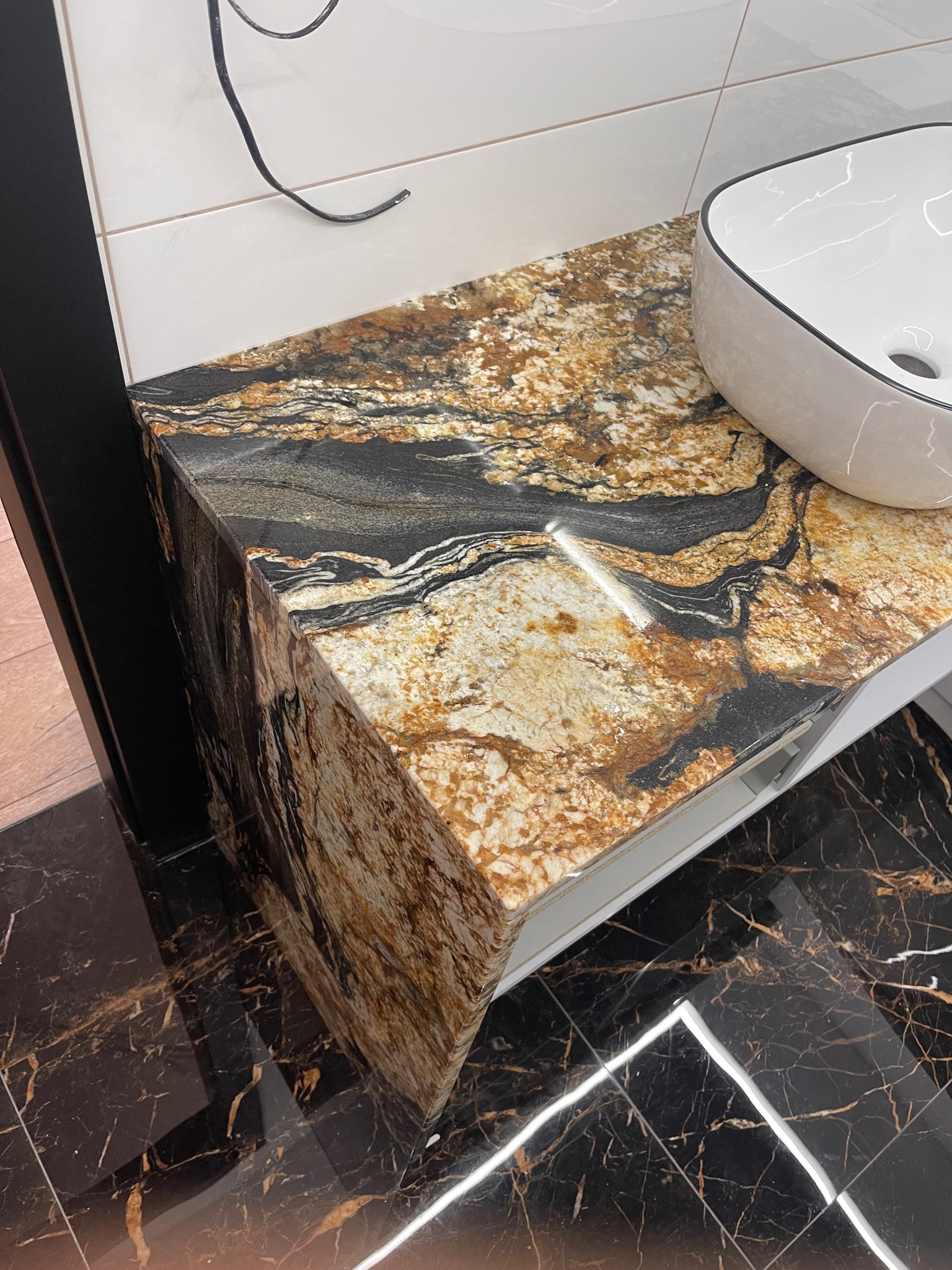 Granitowy blat łazienkowy z umywalką nablatową o wzorzystym wykończeniu, dopasowany do podłogi z ciemnego marmuru.