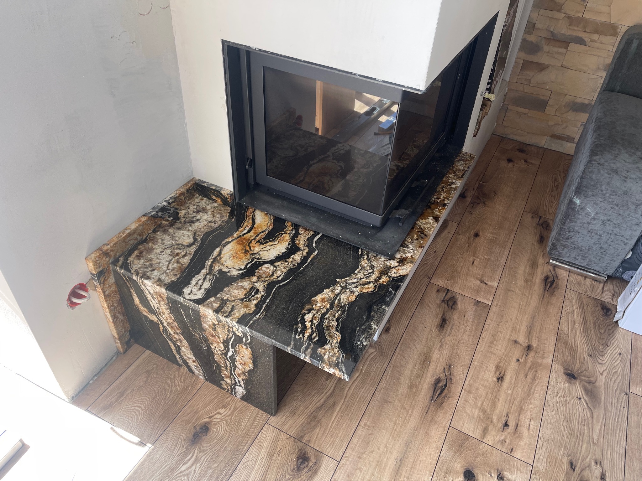 Wykończony fragment kominka z blatem z kamienia o unikalnym wzorze, widoczny na tle drewnianej podłogi i fragmentu ściany. Detal wnętrza.