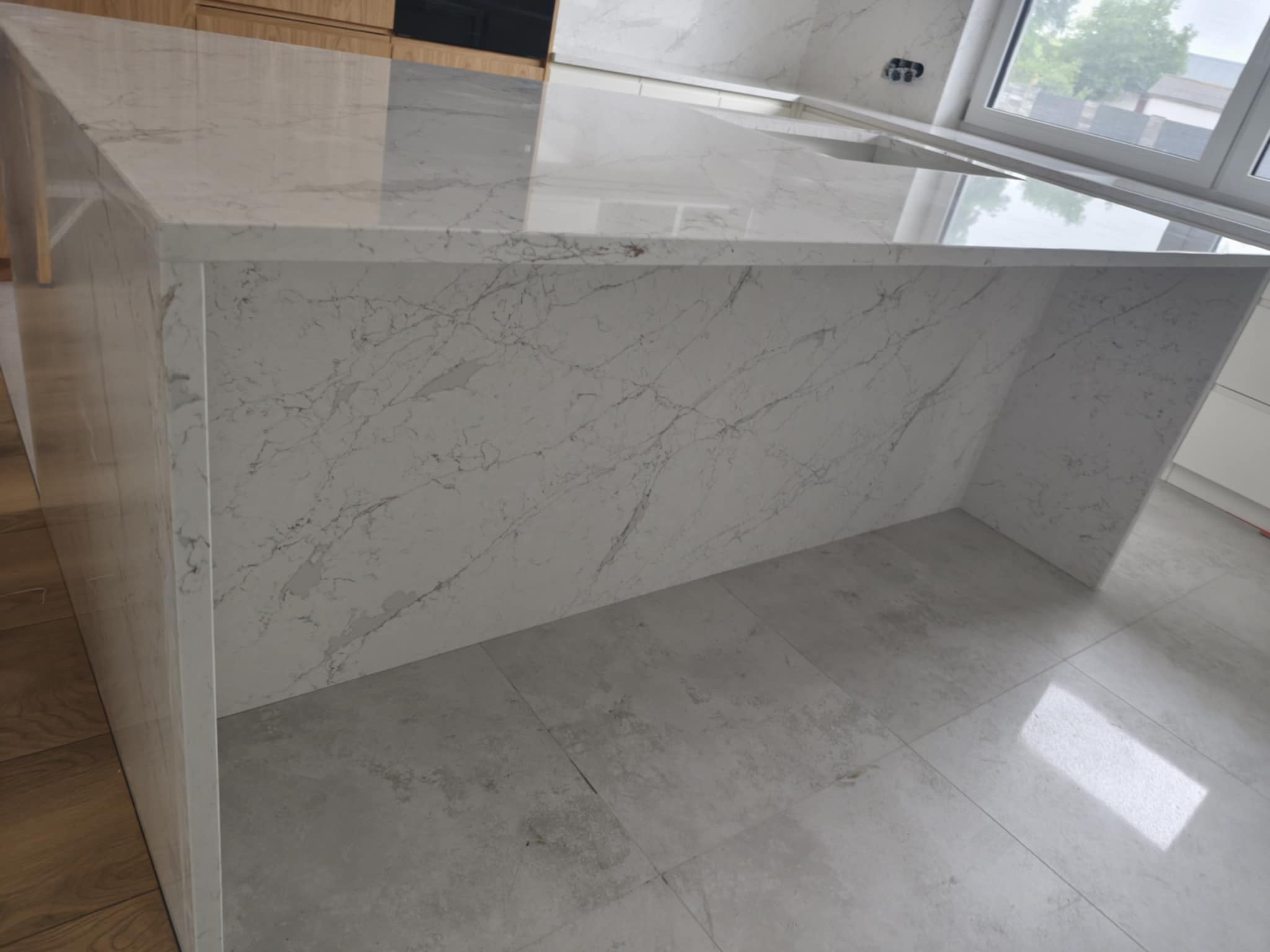 Biały blat kamienny z szarymi żyłkami na wyspie kuchennej, z widoczną strukturą kamienia i odbiciem światła, otoczony jasnymi płytkami podłogowymi.