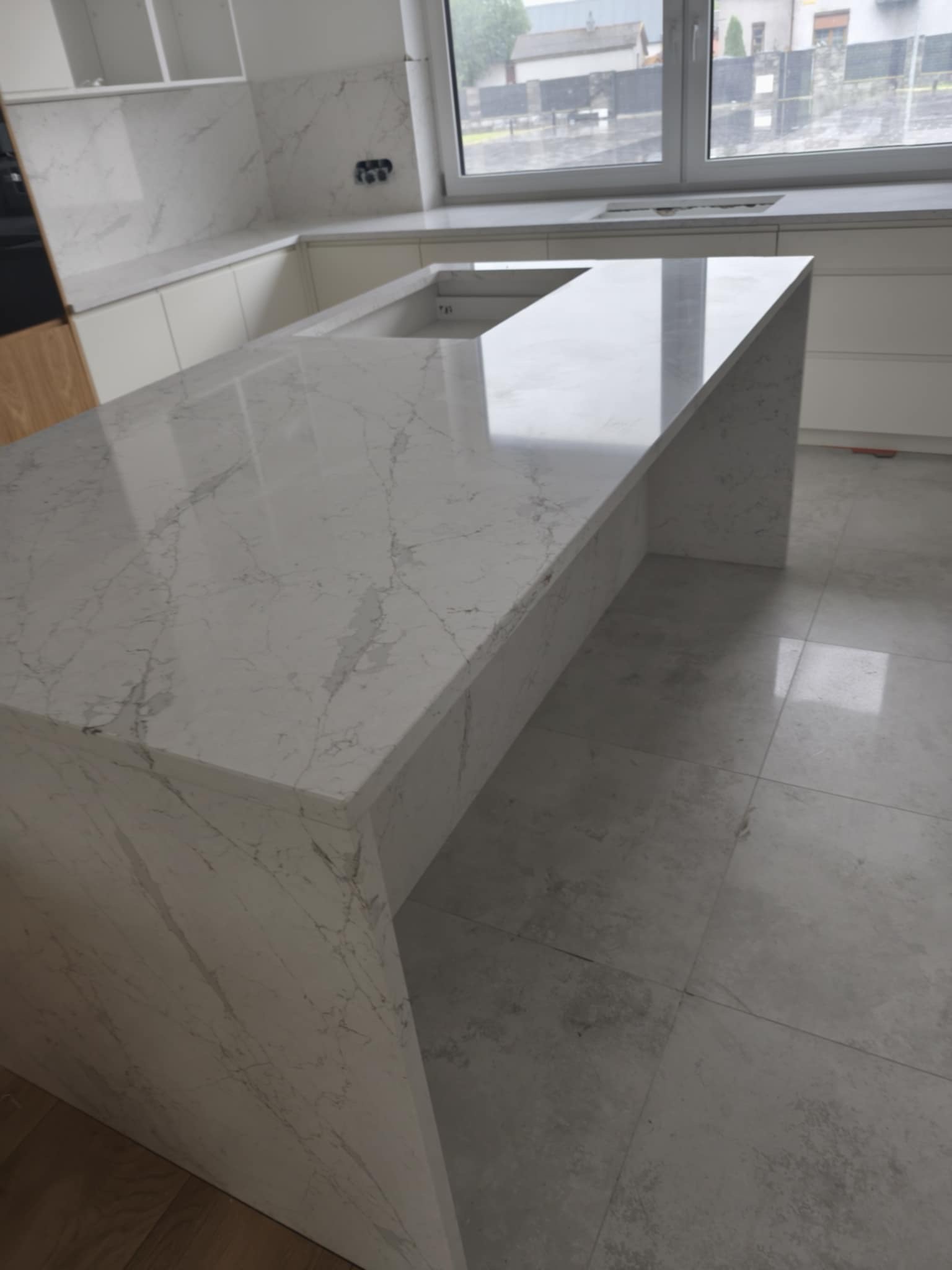 Biały blat kamienny z szarym marmurkowym wzorem, zamontowany w nowoczesnej kuchni z białymi szafkami i dużym oknem w tle. Widoczny zlew w blacie.