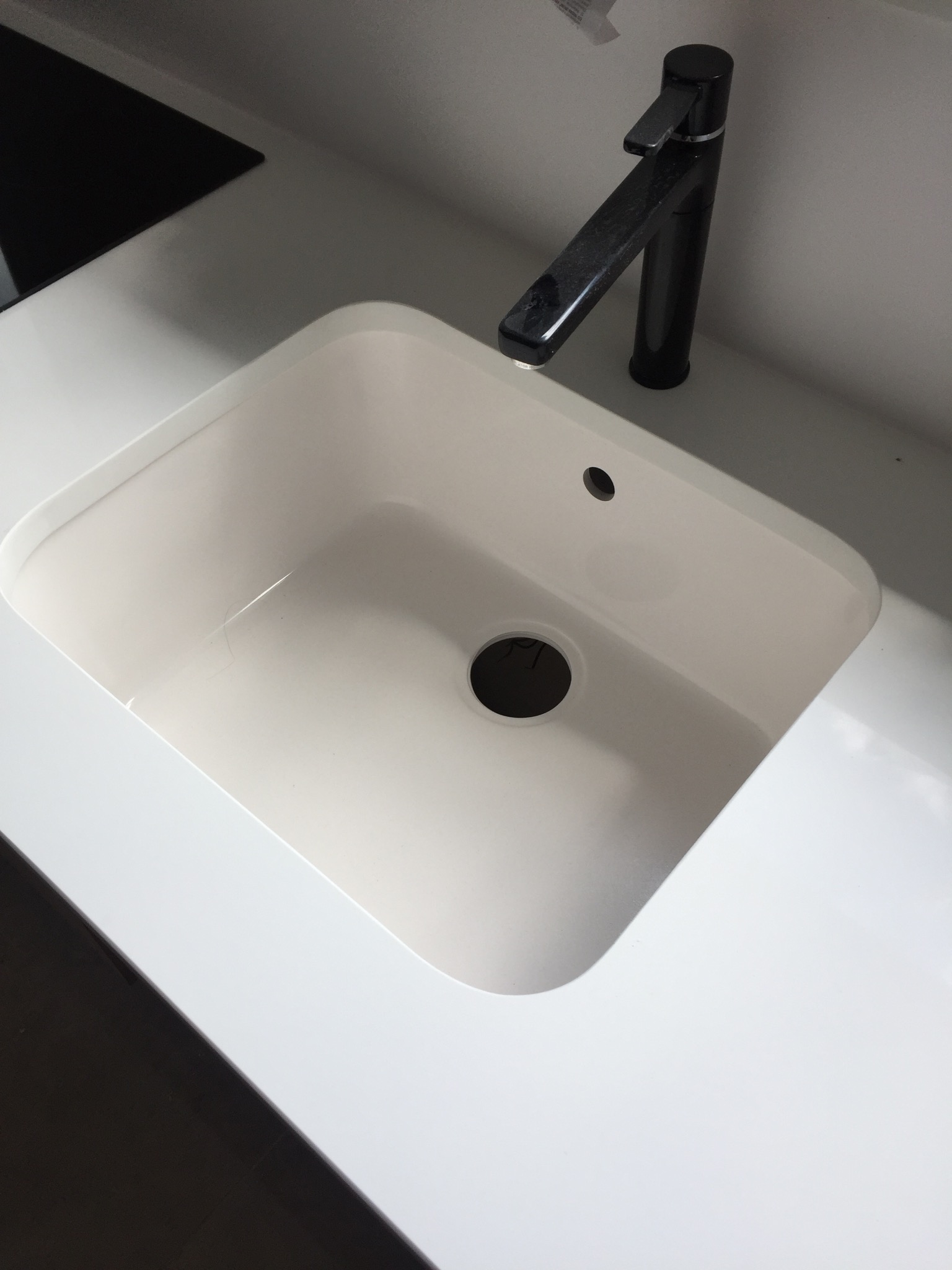 Biały, prostokątny zlew z czarną baterią na blacie, prawdopodobnie kamiennym, z widocznym fragmentem czarnej płyty. Minimalistyczny design.