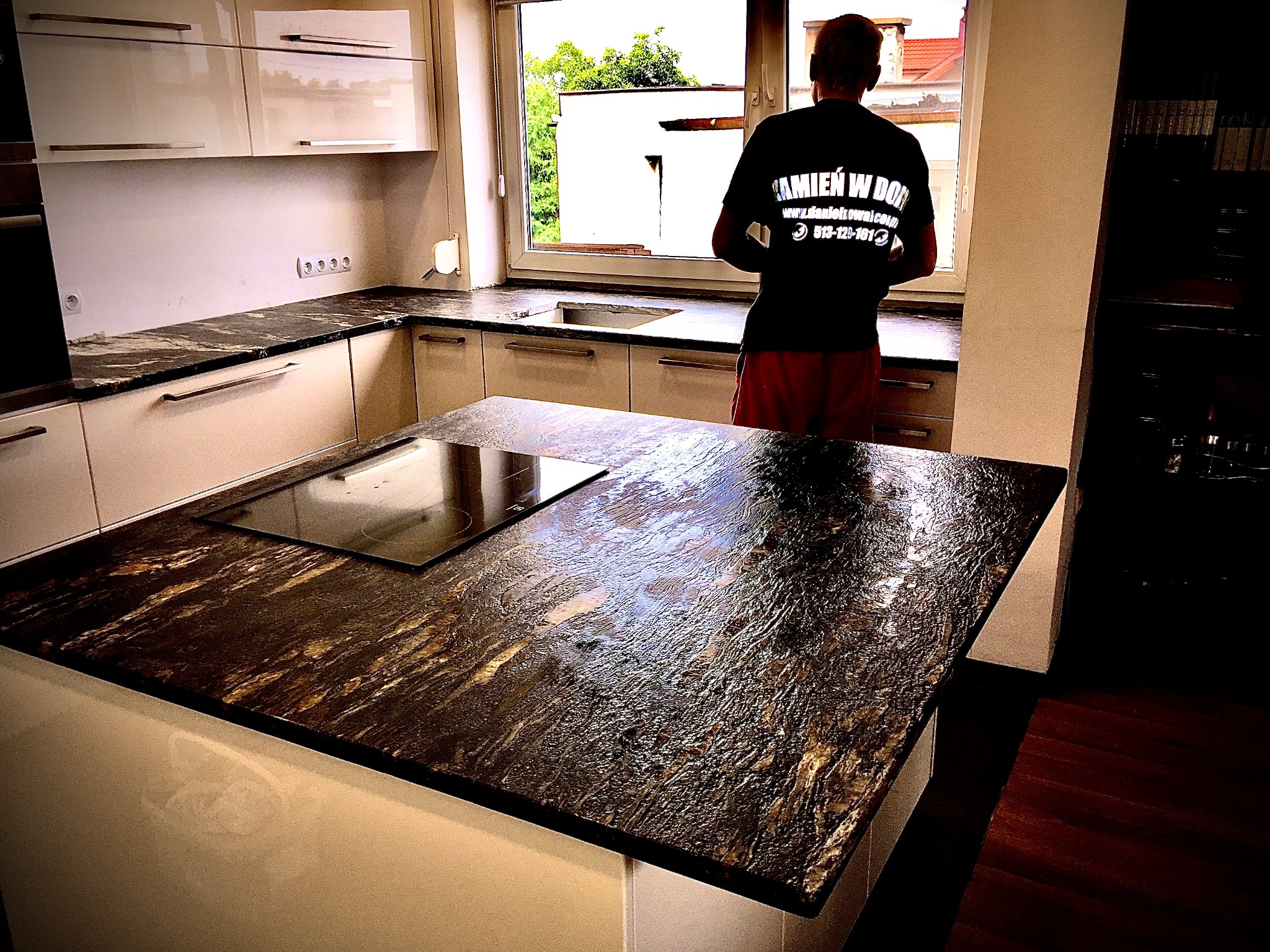 Kuchnia z blatem kamiennym o nieregularnej fakturze, na wyspie płyta indukcyjna. W tle okno i osoba w koszulce z logo firmy kamieniarskiej.