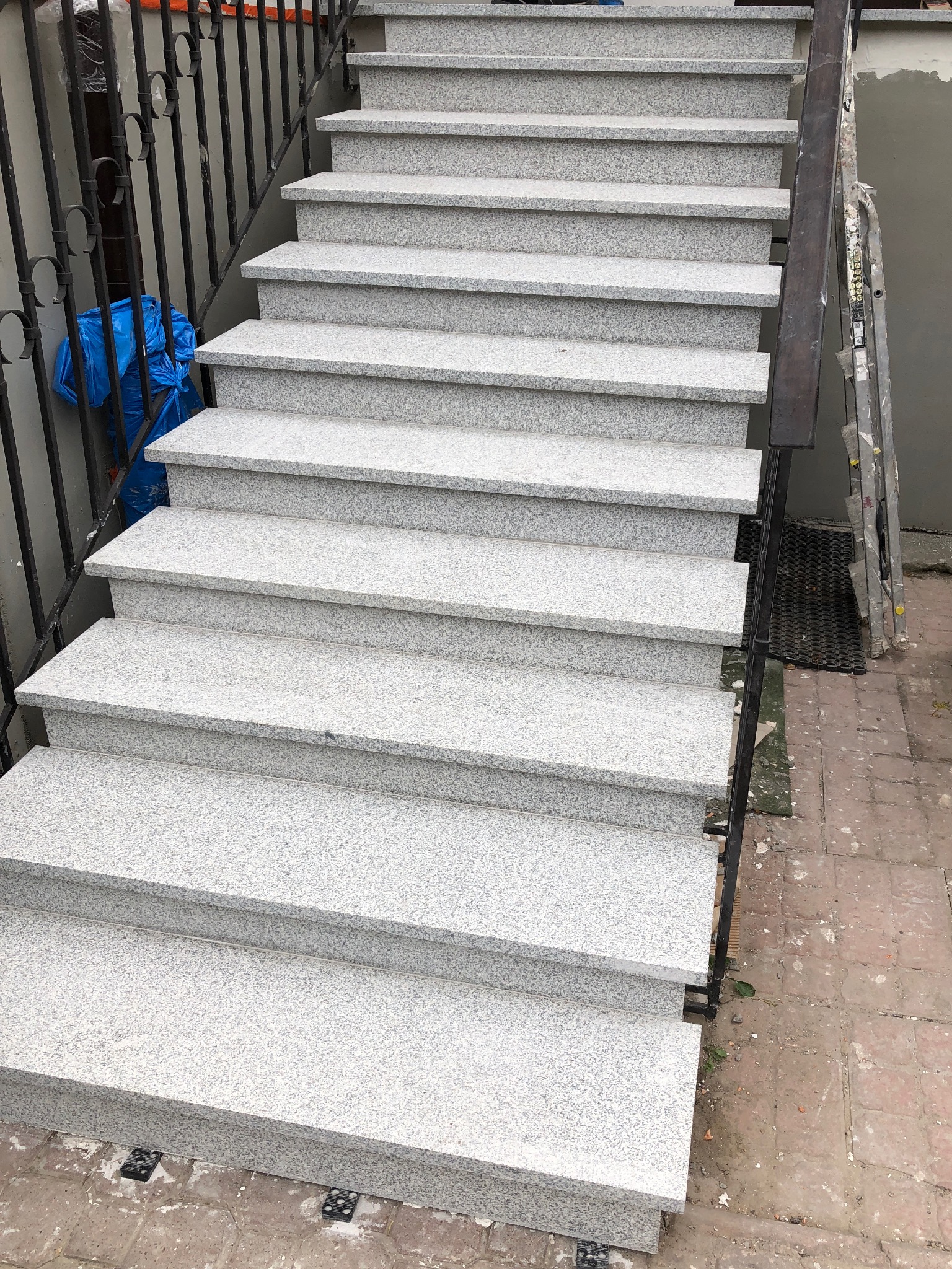 Zewnętrzne schody z jasnego, nakrapianego kamienia z czarną metalową balustradą i drabiną opartą o ścianę w tle. Widok z góry na stopnie.