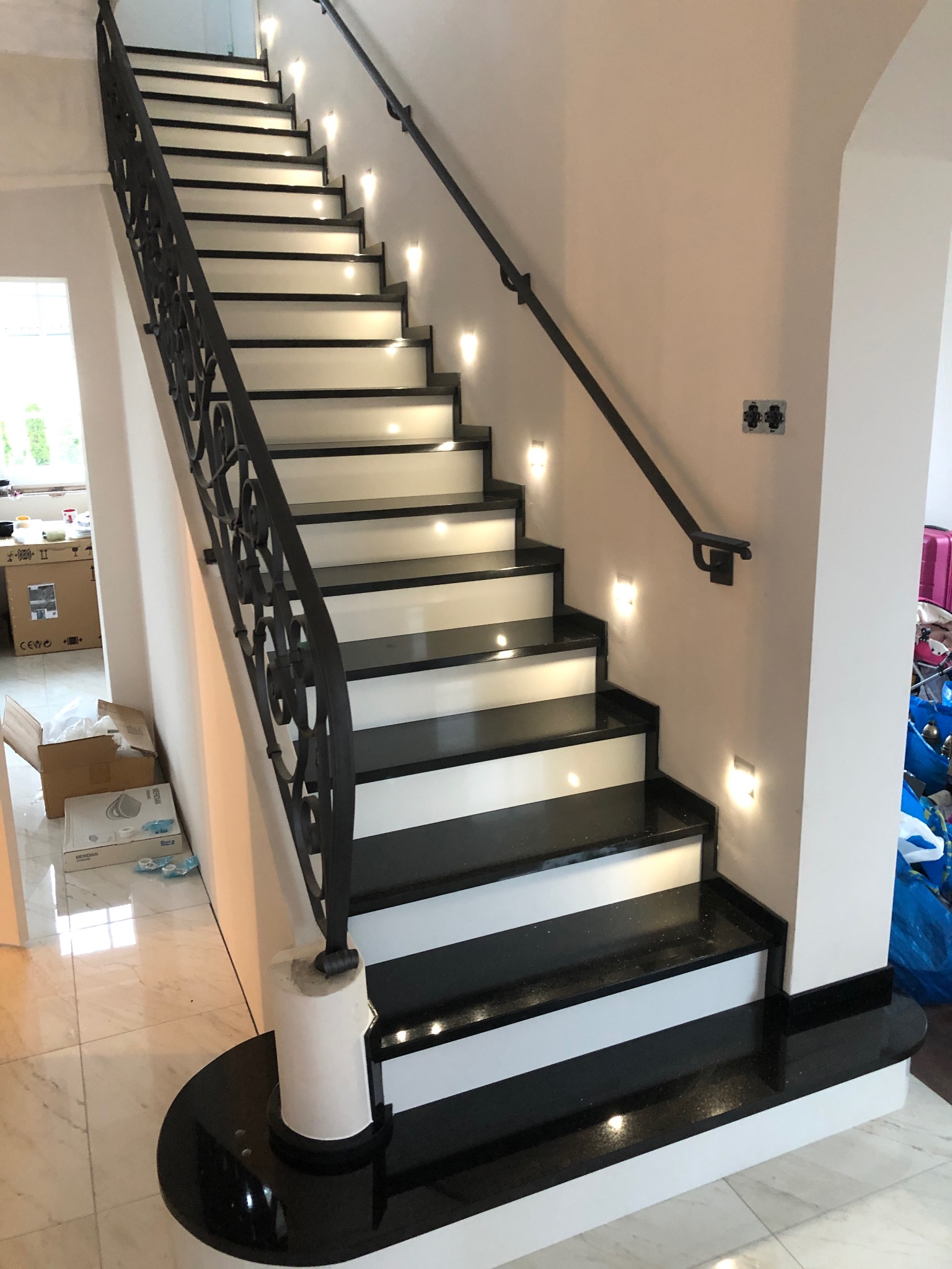 Eleganckie schody z czarnymi kamiennymi stopniami i białymi podstopnicami, oświetlone punktowo, z ozdobną, kutą balustradą i marmurową podłogą.