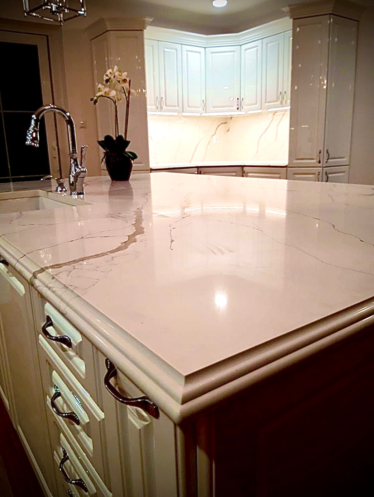 Błyszczący, kremowy blat z subtelnym marmurkowym wzorem na wyspie kuchennej z chromowaną armaturą i białymi szafkami w tle, oświetlony ciepłym światłem.