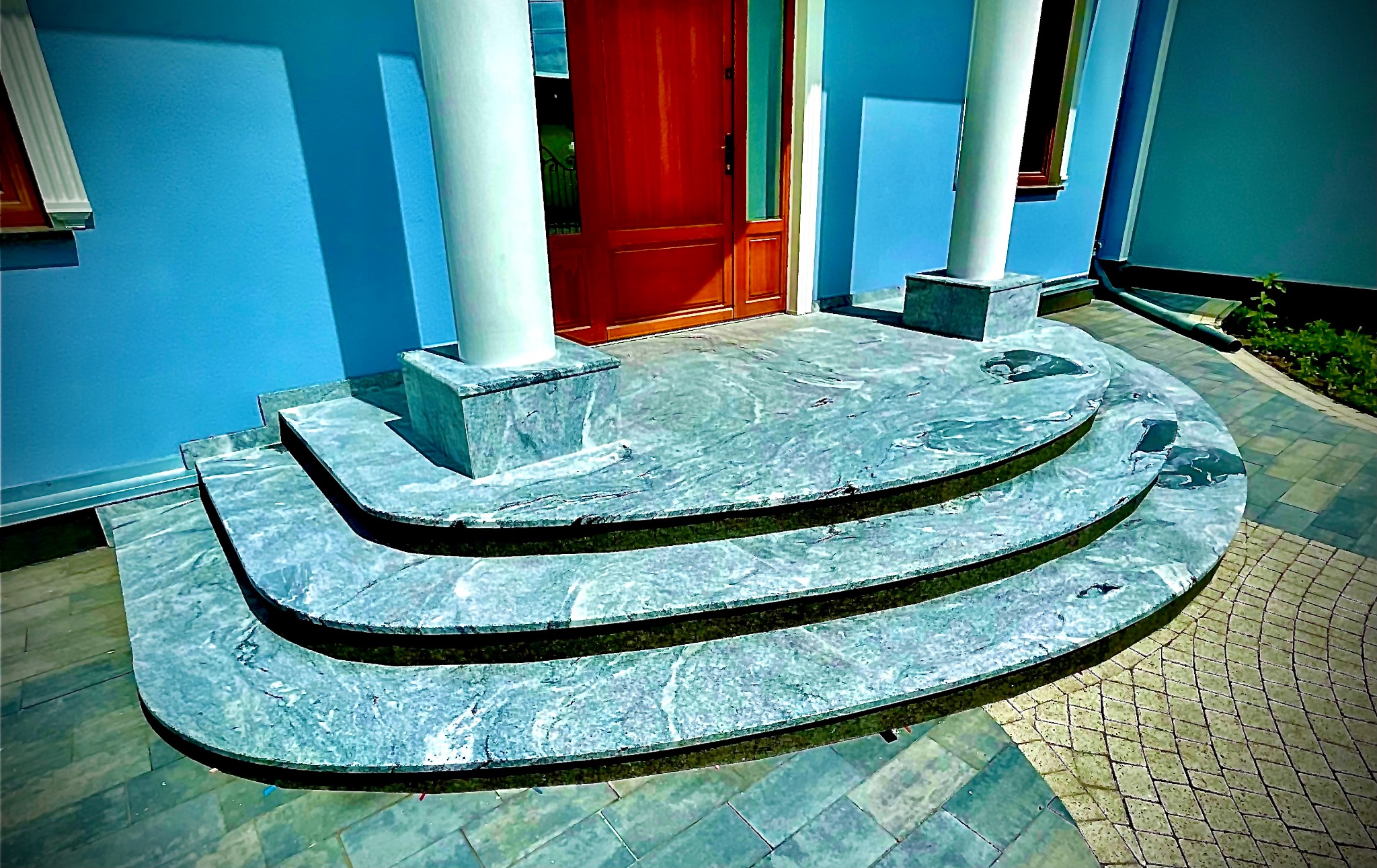 Wejście do domu z trzema marmurowymi stopniami i kolumnami. Niebieska ściana, drewniane drzwi. Widok na schody i podjazd z kostki brukowej.