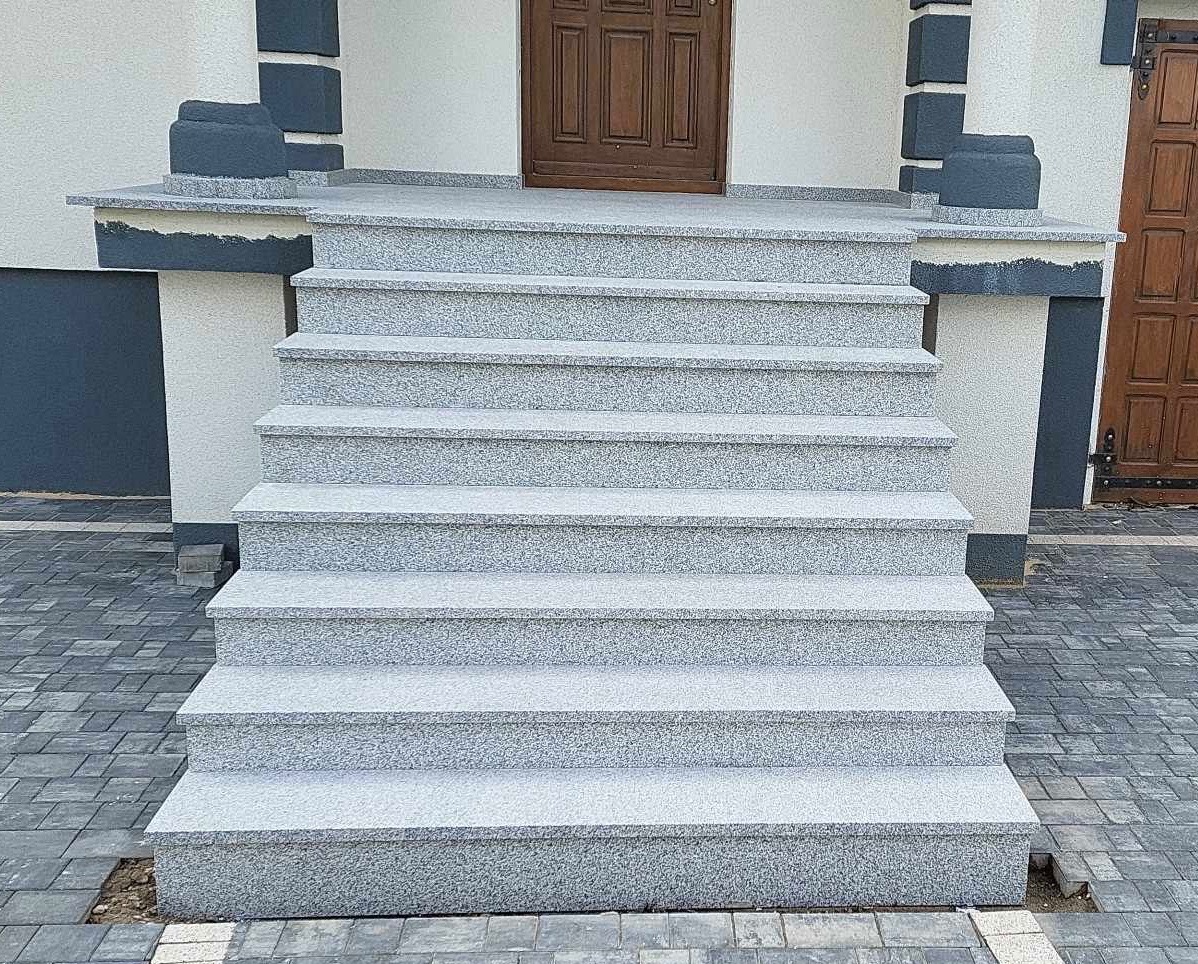 Zewnętrzne schody z jasnoszarego kamienia prowadzące do drzwi wejściowych, z kostką brukową w odcieniach szarości u podstawy. Detale architektoniczne w kolorze grafitowym.