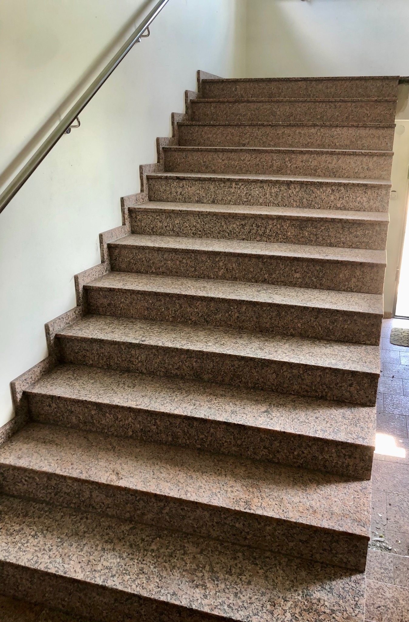 Kamienne schody wewnętrzne z metalową barierką, widok z dołu. Stopnie wykonane z brązowego kamienia z drobnym wzorem, jasne ściany, fragment podłogi i drzwi.