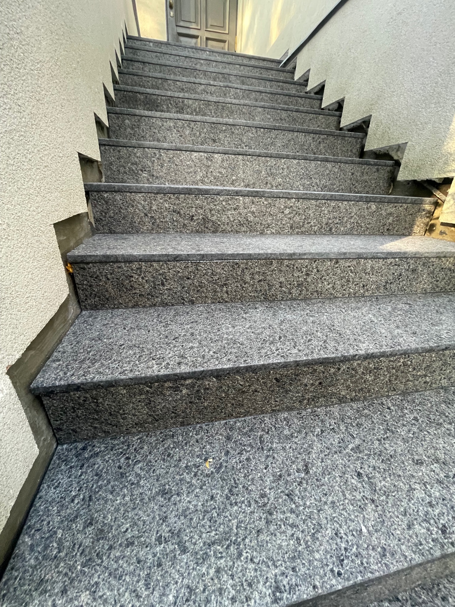 Zewnętrzne schody z szarego kamienia prowadzą w górę, widok z dołu. Ściana z szorstkim tynkiem po lewej stronie. Górne stopnie oświetlone słońcem.