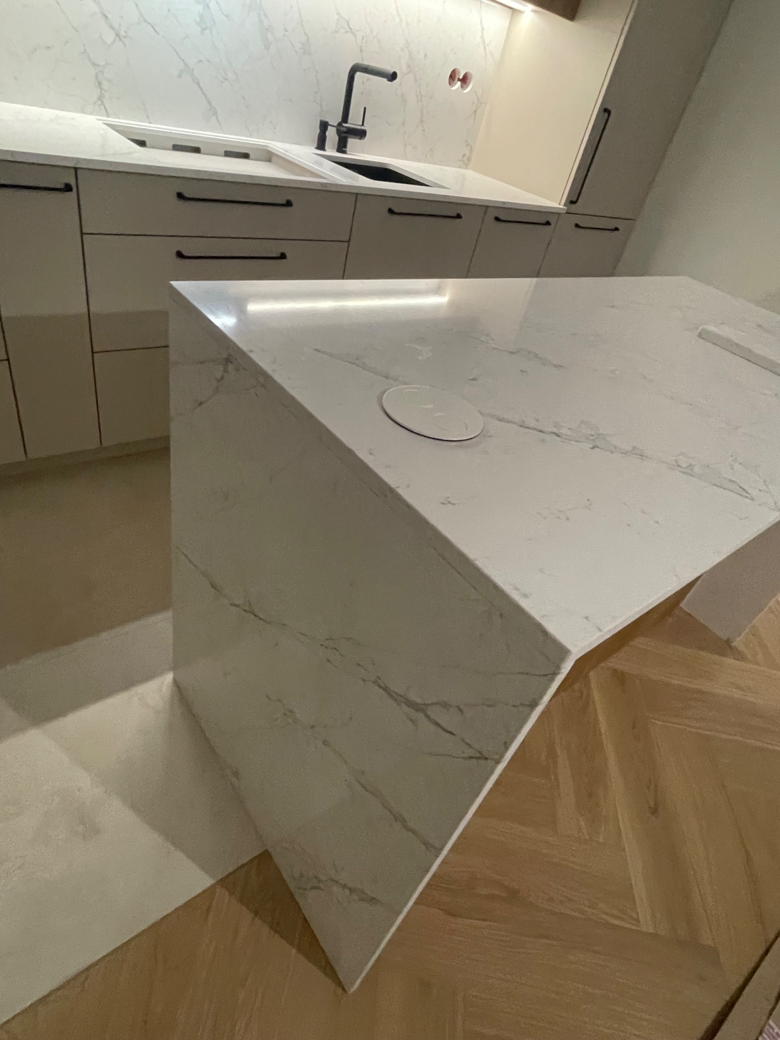 Biały blat kamienny z marmurowym wzorem na wyspie kuchennej, z widocznym gniazdkiem elektrycznym i fragmentem parkietu ułożonego w jodełkę.