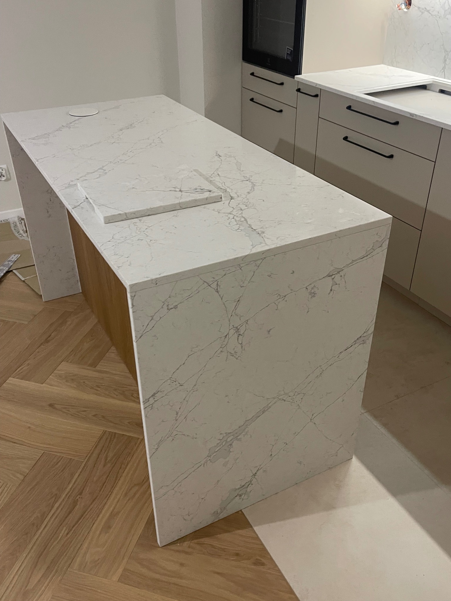 Biały blat kamienny z marmurkowym wzorem, tworzący wyspę kuchenną z drewnianym wykończeniem. Widoczne gniazdko i fragment zlewu. W tle zabudowa kuchenna.