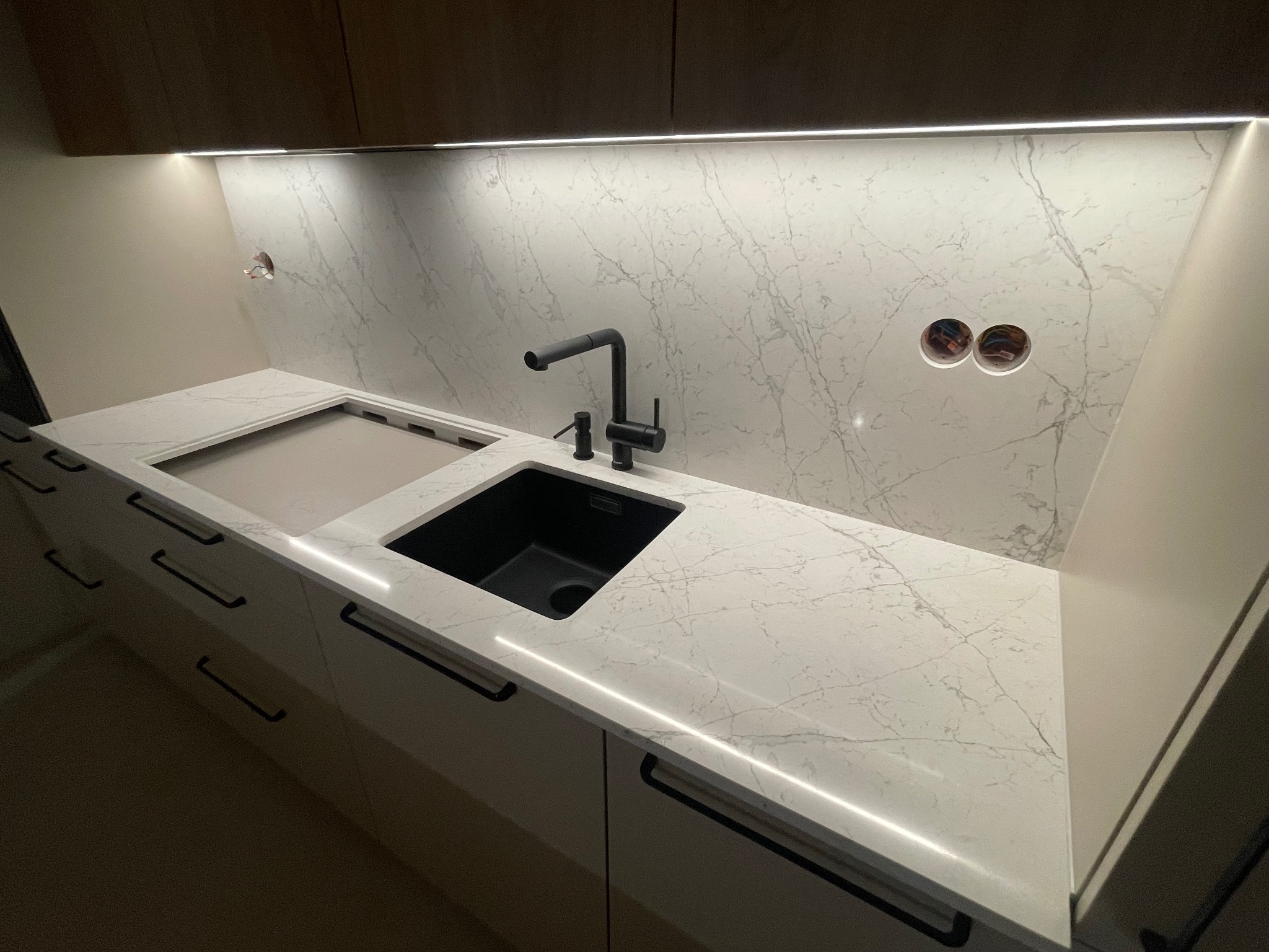 Biały blat kamienny z czarnym zlewem i baterią w nowoczesnej kuchni, podświetlony LED, z marmurkowym wzorem na ścianie i widocznymi gniazdkami elektrycznymi.