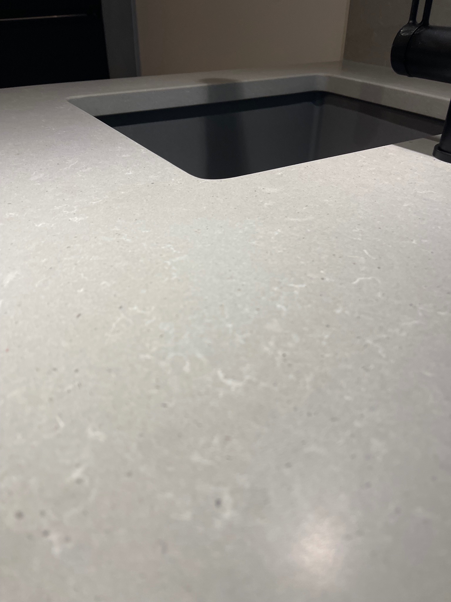 Jasnoszary blat kamienny z wyciętym otworem na zlew, widoczny fragment czarnej baterii. Delikatna faktura kamienia, minimalistyczny design.