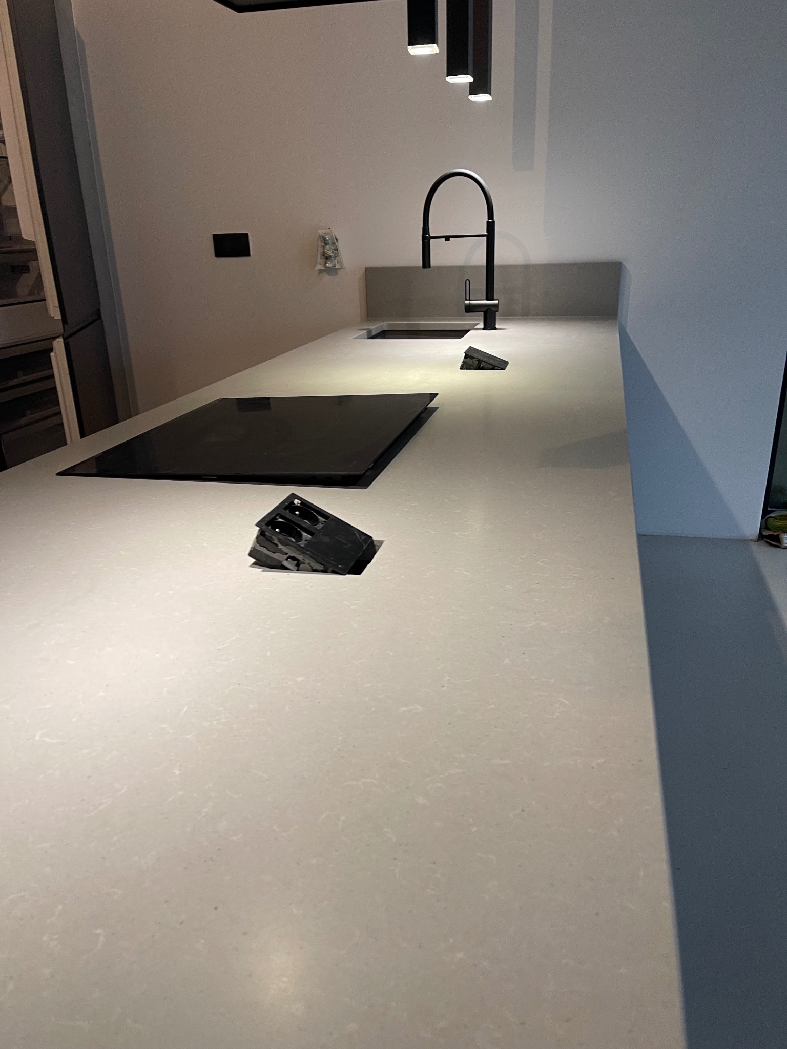 Jasny blat kamienny z czarną płytą indukcyjną i minimalistyczną baterią kuchenną. Nowoczesny design, subtelne oświetlenie punktowe, widoczny organizer na noże.