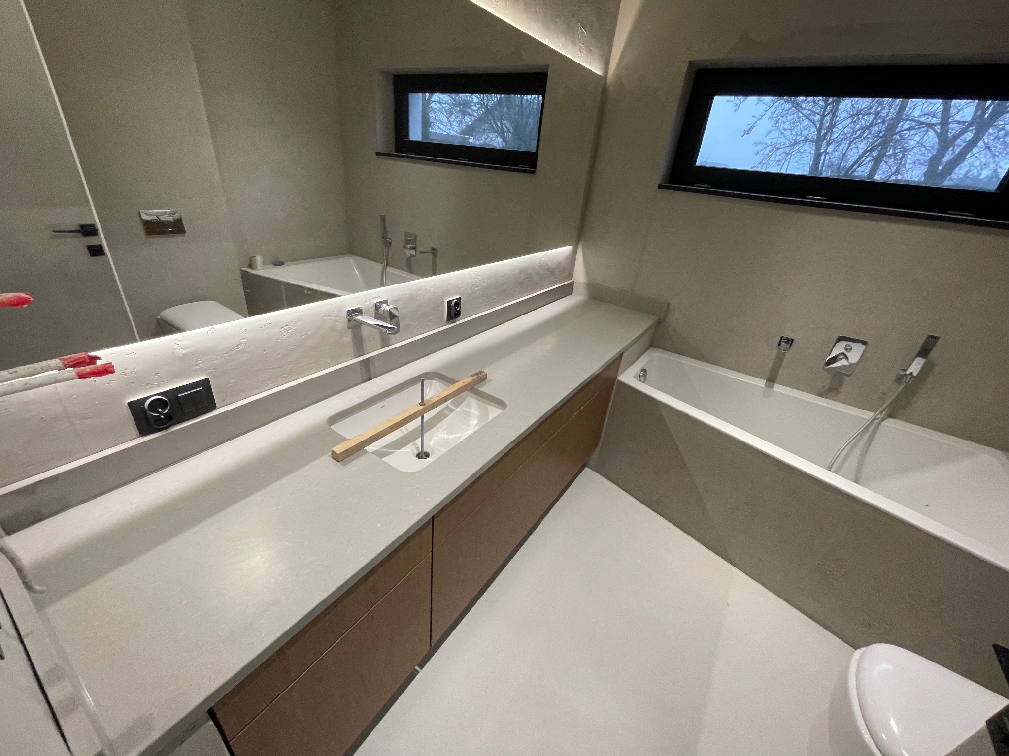 Jasny blat kamienny w nowoczesnej łazience z drewnianymi szafkami, oświetleniem LED i widokiem z okna na drzewa. Minimalistyczny design, neutralna kolorystyka.