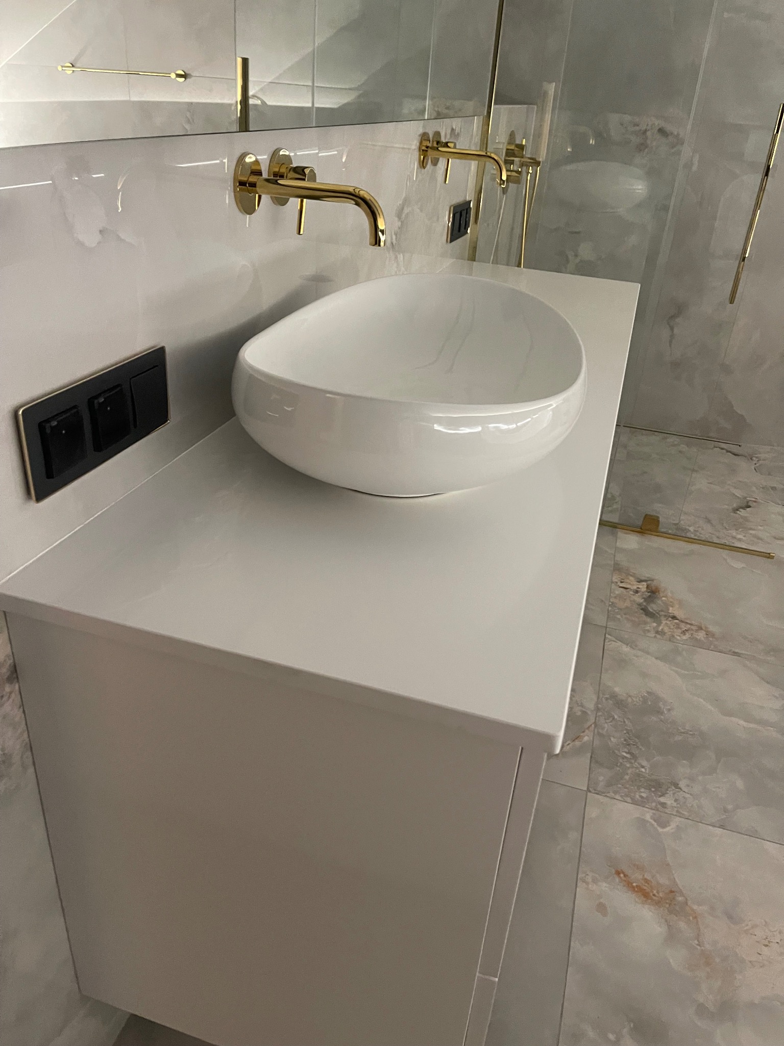 Biała, owalna umywalka nablatowa z baterią w kolorze złotym, zamontowana na szafce z blatem. W tle marmurowe płytki i szklana kabina prysznicowa.