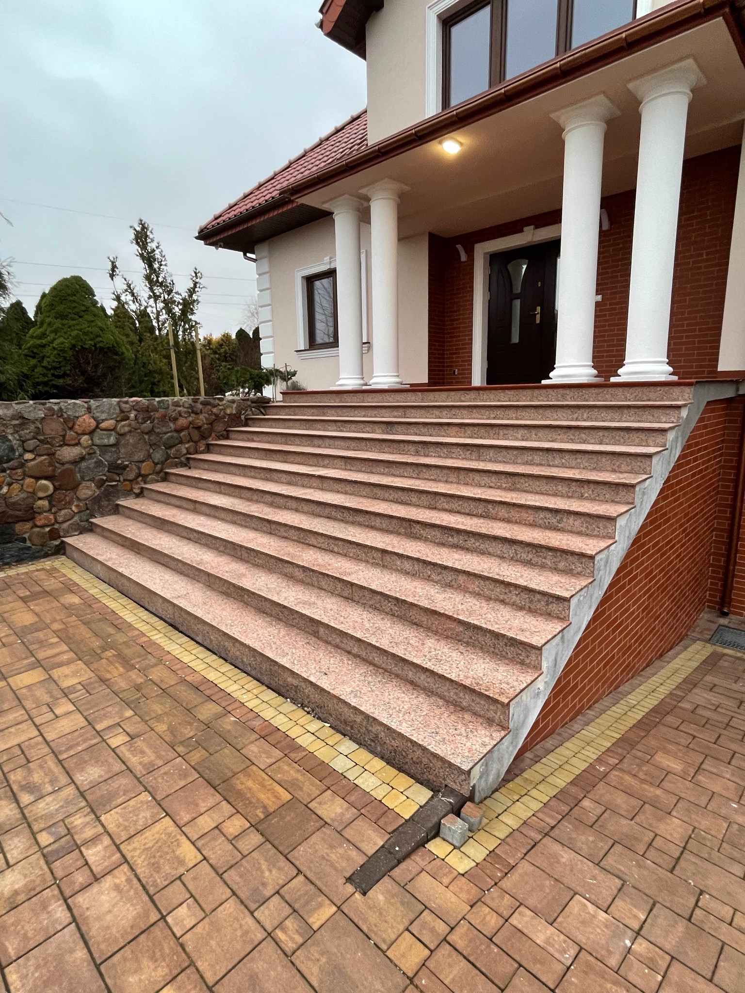 Zewnętrzne schody z różowego kamienia prowadzące do domu z kolumnami, otoczone brukowaną ścieżką i kamiennym murem. Pochmurne niebo w tle.
