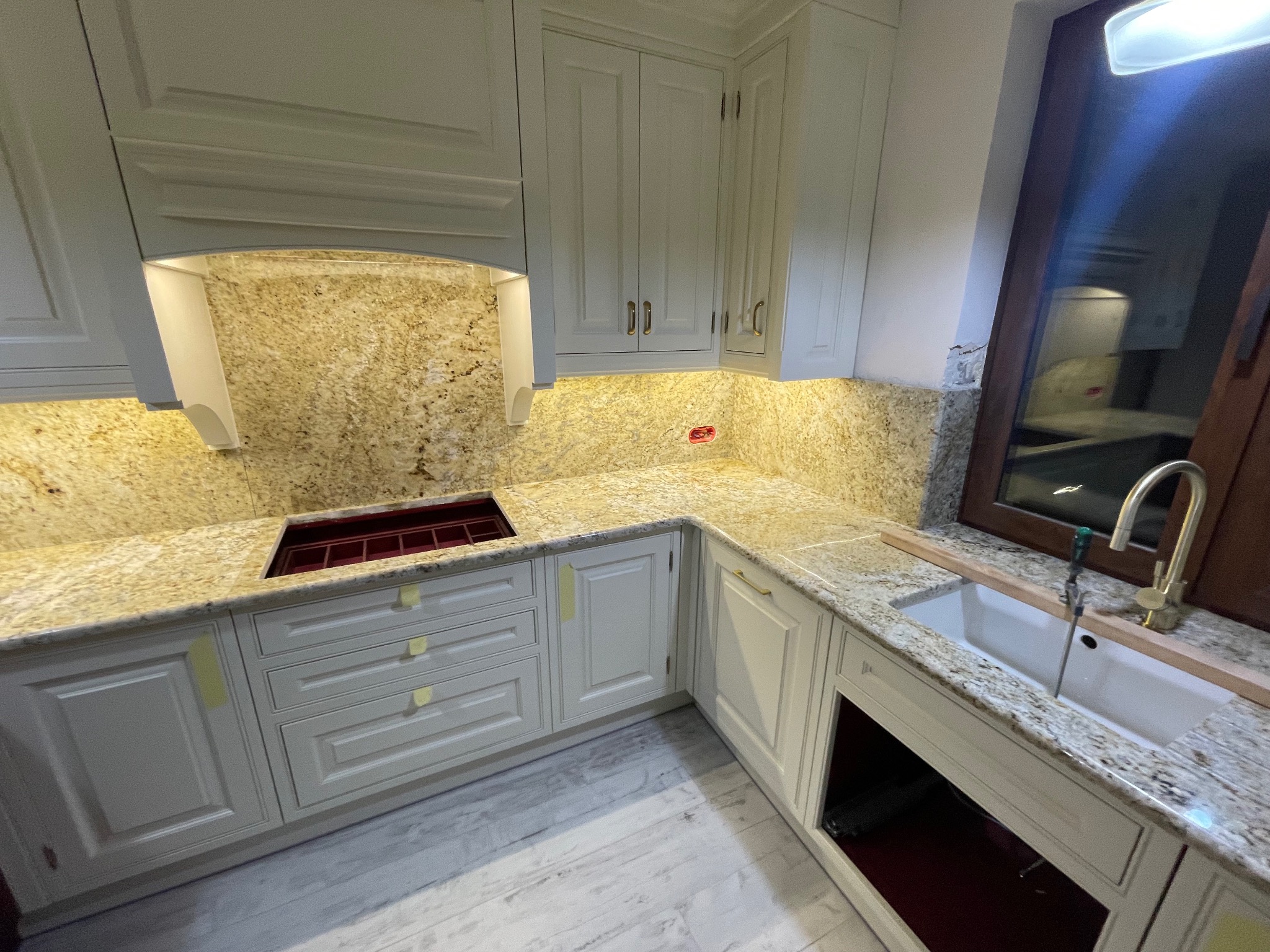 Kuchnia z blatem z kamienia naturalnego w odcieniach beżu i brązu, białe szafki z frezowanymi frontami, widoczny zlew i bateria. Oświetlenie podszafkowe.